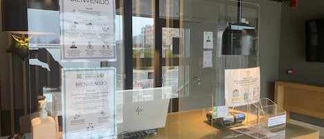 A reception desk with a laptop, a glass partition, and COVID-19 safety information signs.