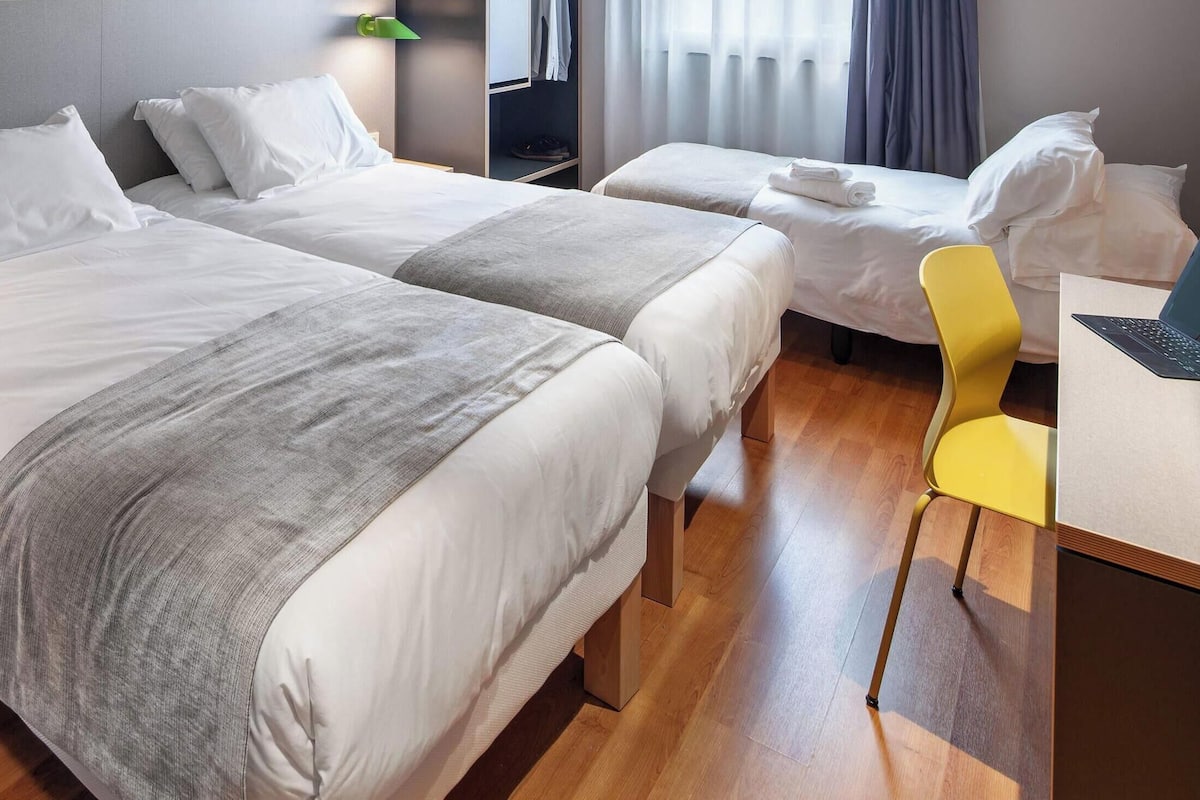 A hotel room with two beds, a desk, a chair, and a laptop.