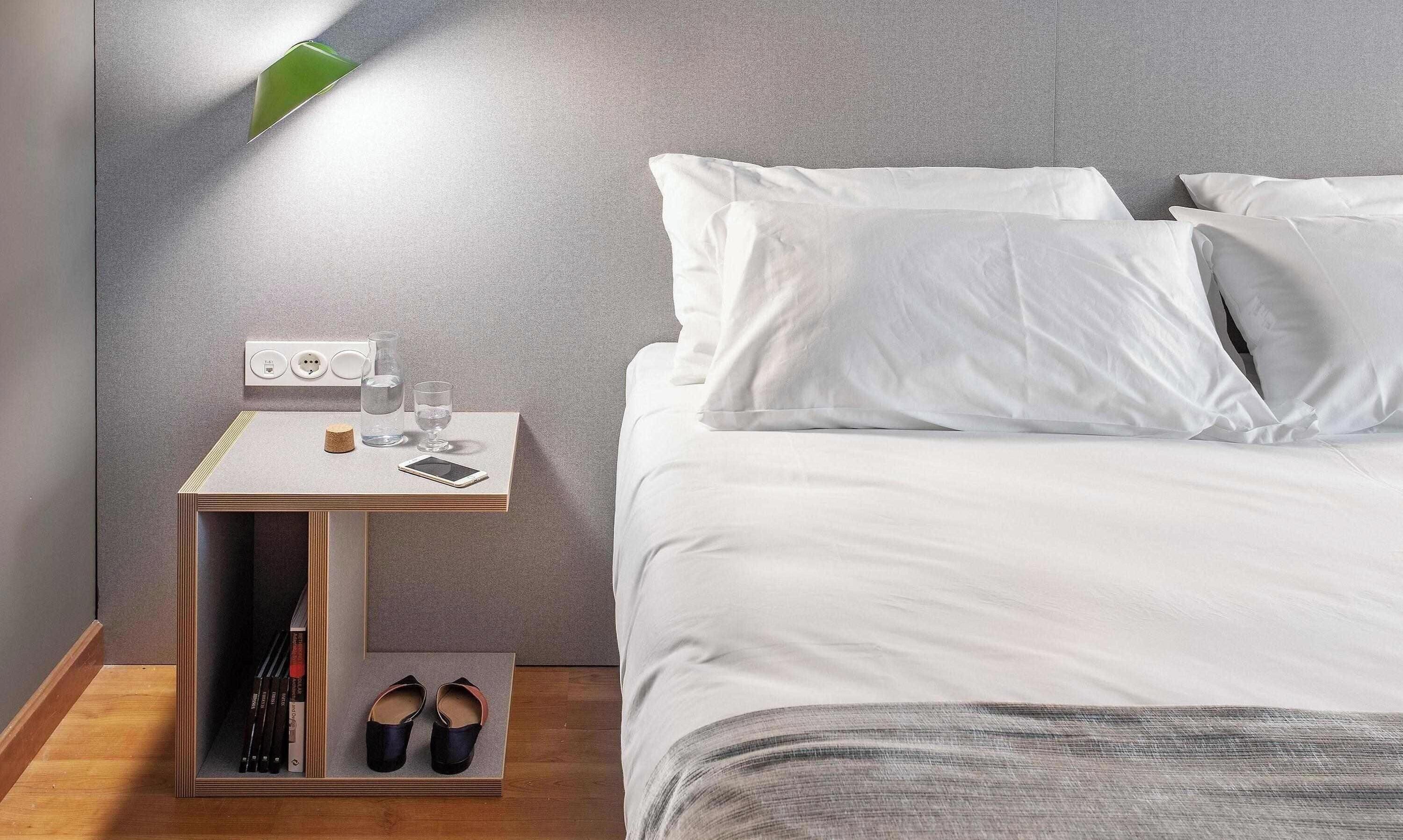 Ein ordentlich bezogenes Bett mit weißen Leinen, ein Nachttisch mit Lampe, ein Glas Wasser, ein Smartphone und ein Paar Schuhe auf dem Boden.