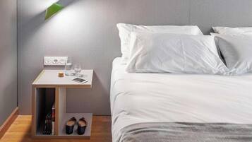 Una cama bien hecha con sábanas blancas, una mesita de noche con lámpara, un vaso de agua, un teléfono inteligente y un par de zapatos en el suelo.