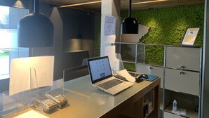 Un espacio de oficina moderno con un escritorio de vidrio, una computadora portátil, un teléfono móvil y una computadora de escritorio. Hay una pared verde con un diseño texturizado, una lámpara colgante y un archivador de metal.