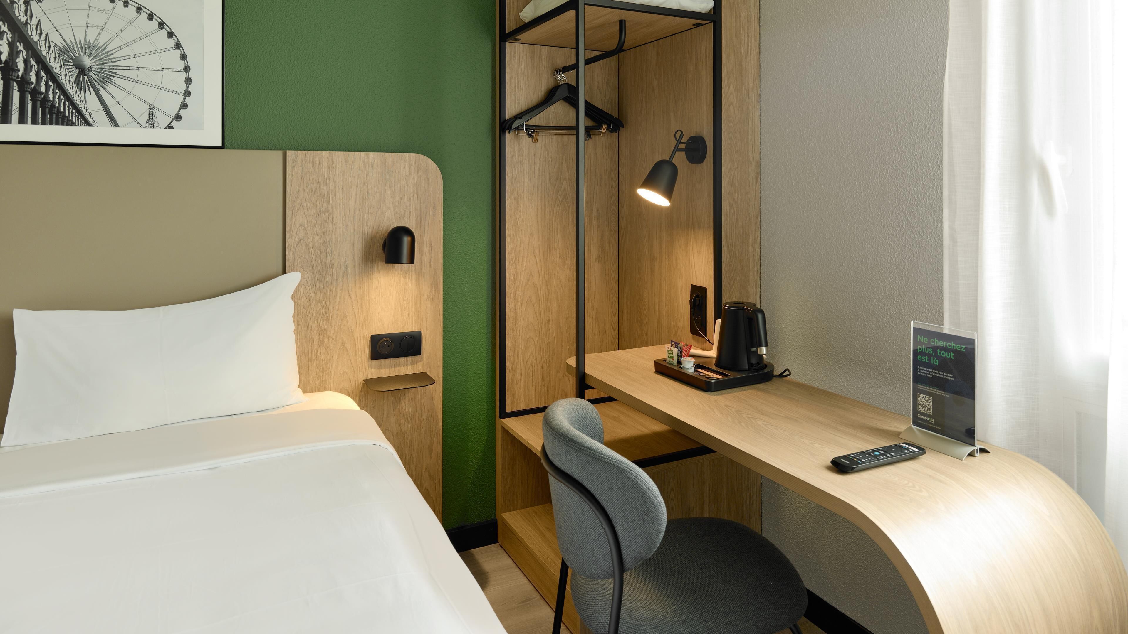 A hotel room with a bed, a desk with a chair, a coffee maker, a hanging light, and a framed picture on the wall.