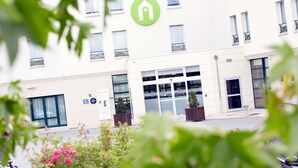 Entrada de hotel com logotipo verde, grandes janelas e placa indicando os serviços.