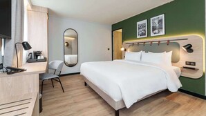 Quarto de hotel moderno com uma cama grande, uma escrivaninha com cadeira, um espelho e uma televisão.