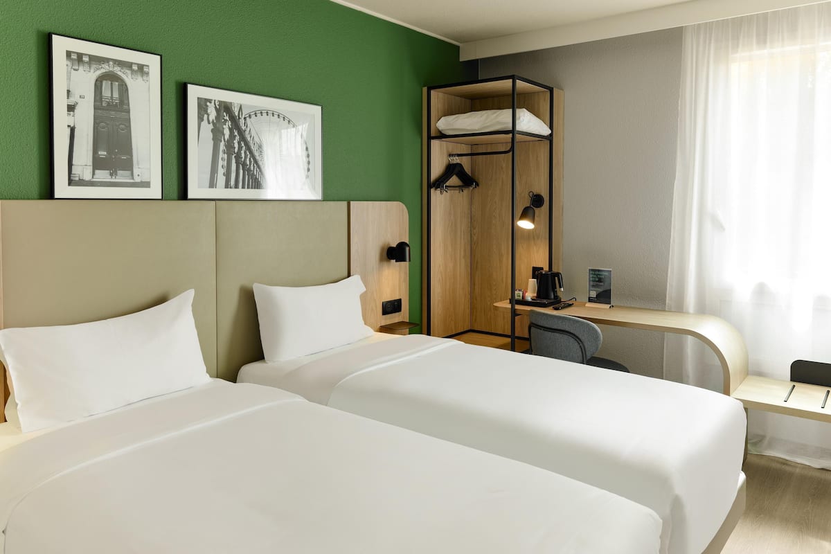 Ein Hotelzimmer mit zwei Betten, einem Schreibtisch, einem Stuhl, einer Duschkabine und zwei gerahmten Bildern an der Wand.