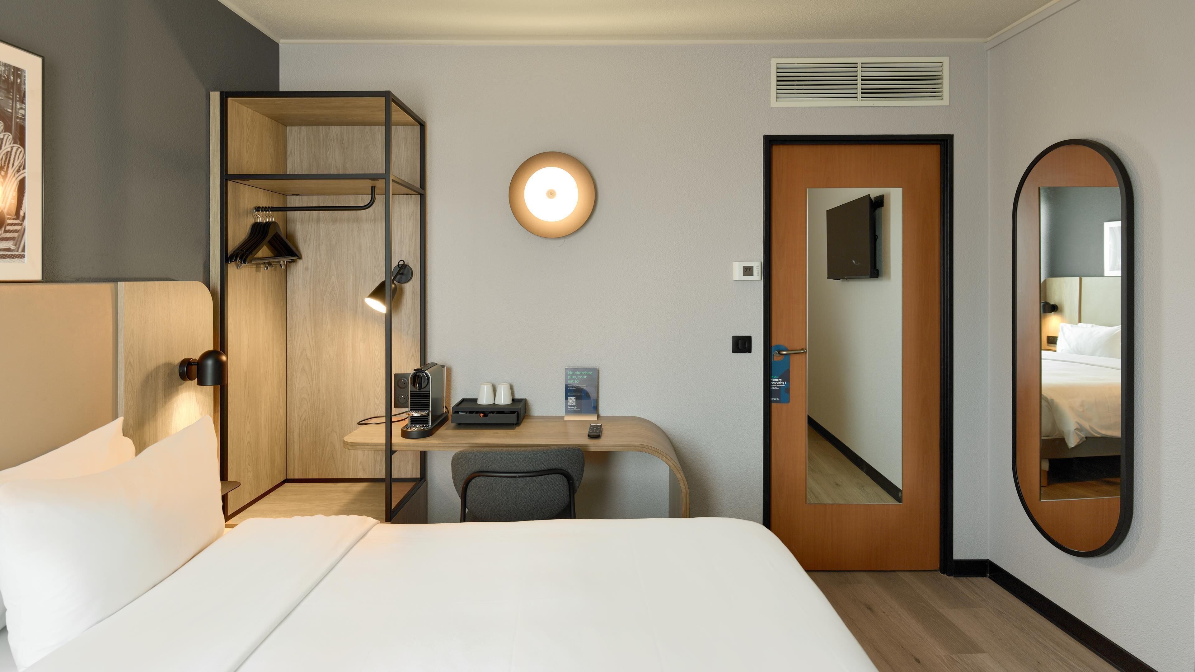 Quarto de hotel com cama, escrivaninha com luminária, espelho, guarda-roupa e luz acoplada na parede.