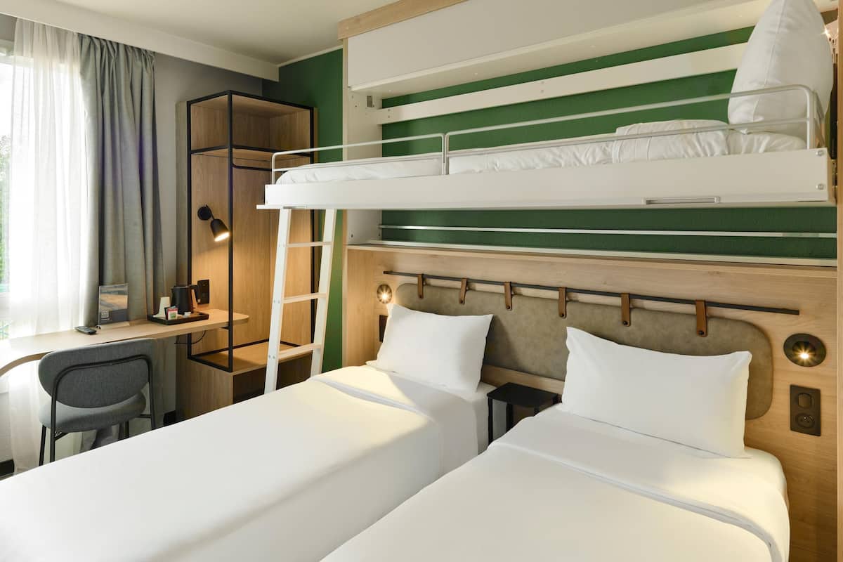 Ein Hotelzimmer mit einem Etagenbett, einem Schreibtisch mit Stuhl, einem kleinen Tisch, einer Lampe und einem Fenster mit Vorhängen.