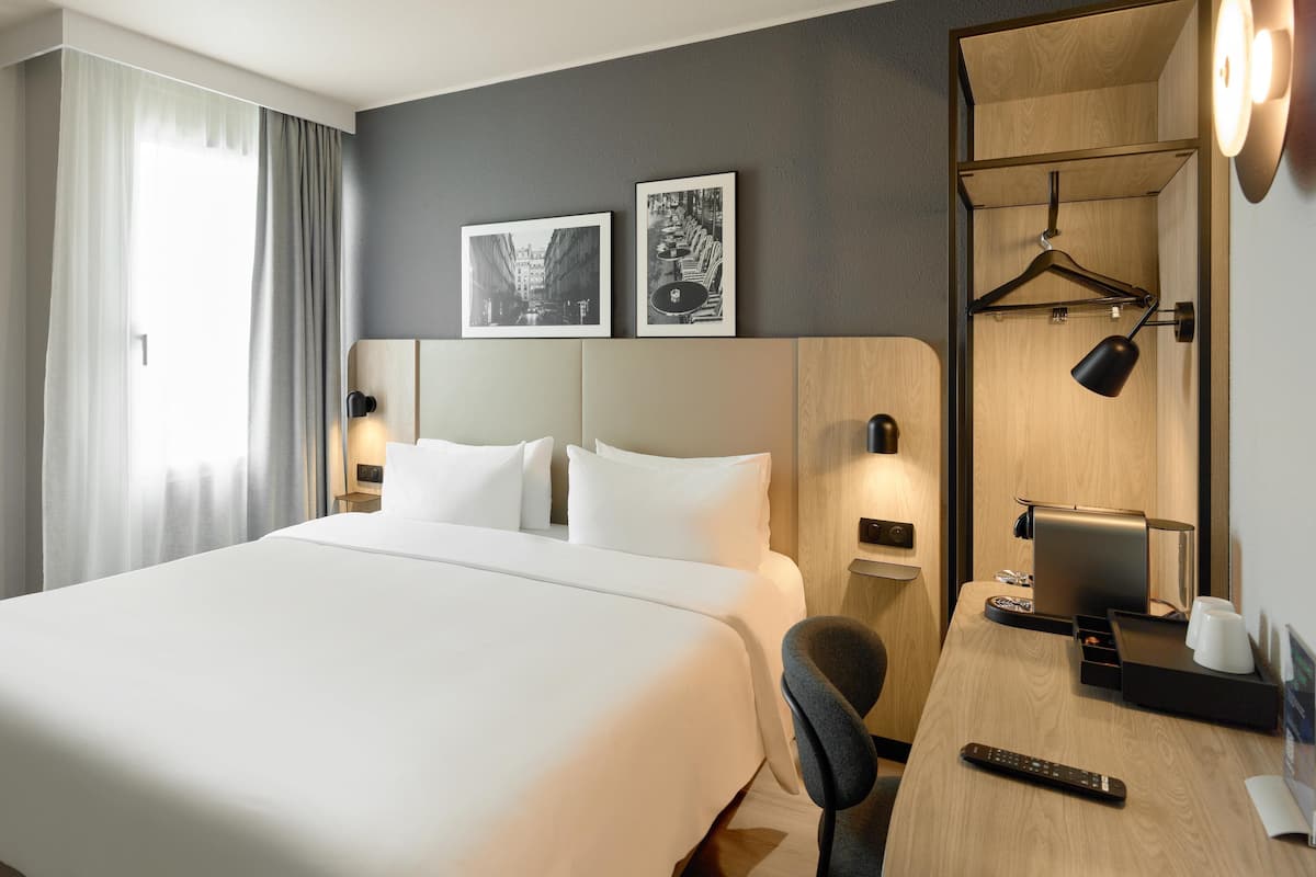 Ein modernes Hotelzimmer mit einem großen Bett, einem Schreibtisch mit Kaffeemaschine, einem Sessel, einer Schreibtischlampe und zwei gerahmten Bildern an der Wand.