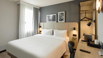 Una habitación de hotel con una cama grande, un escritorio con cafetera, una silla, una lámpara de escritorio y dos cuadros en la pared.