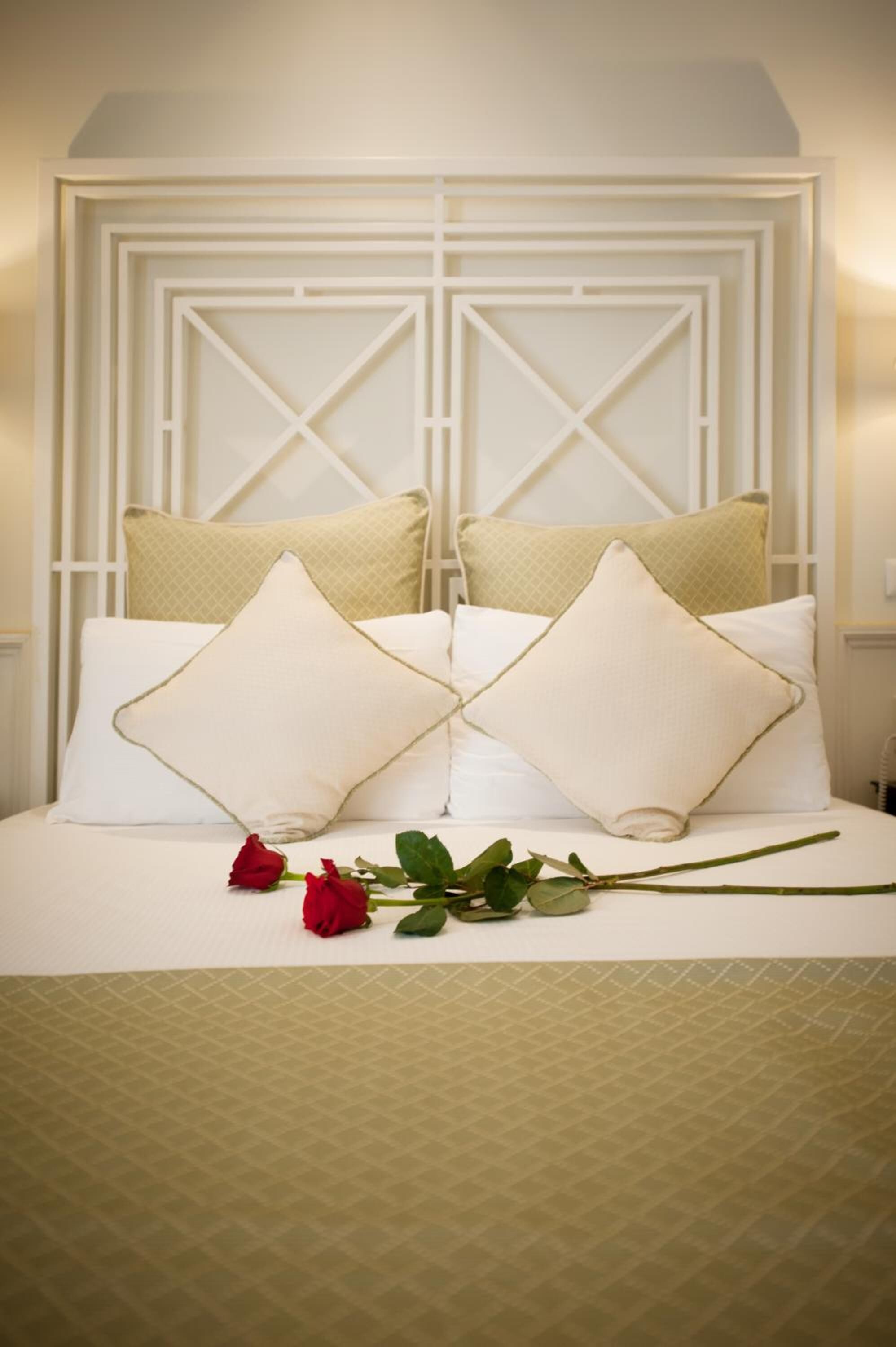 A neatly made bed with white bedding and two pillows, a red rose, and a green branch on the bed.