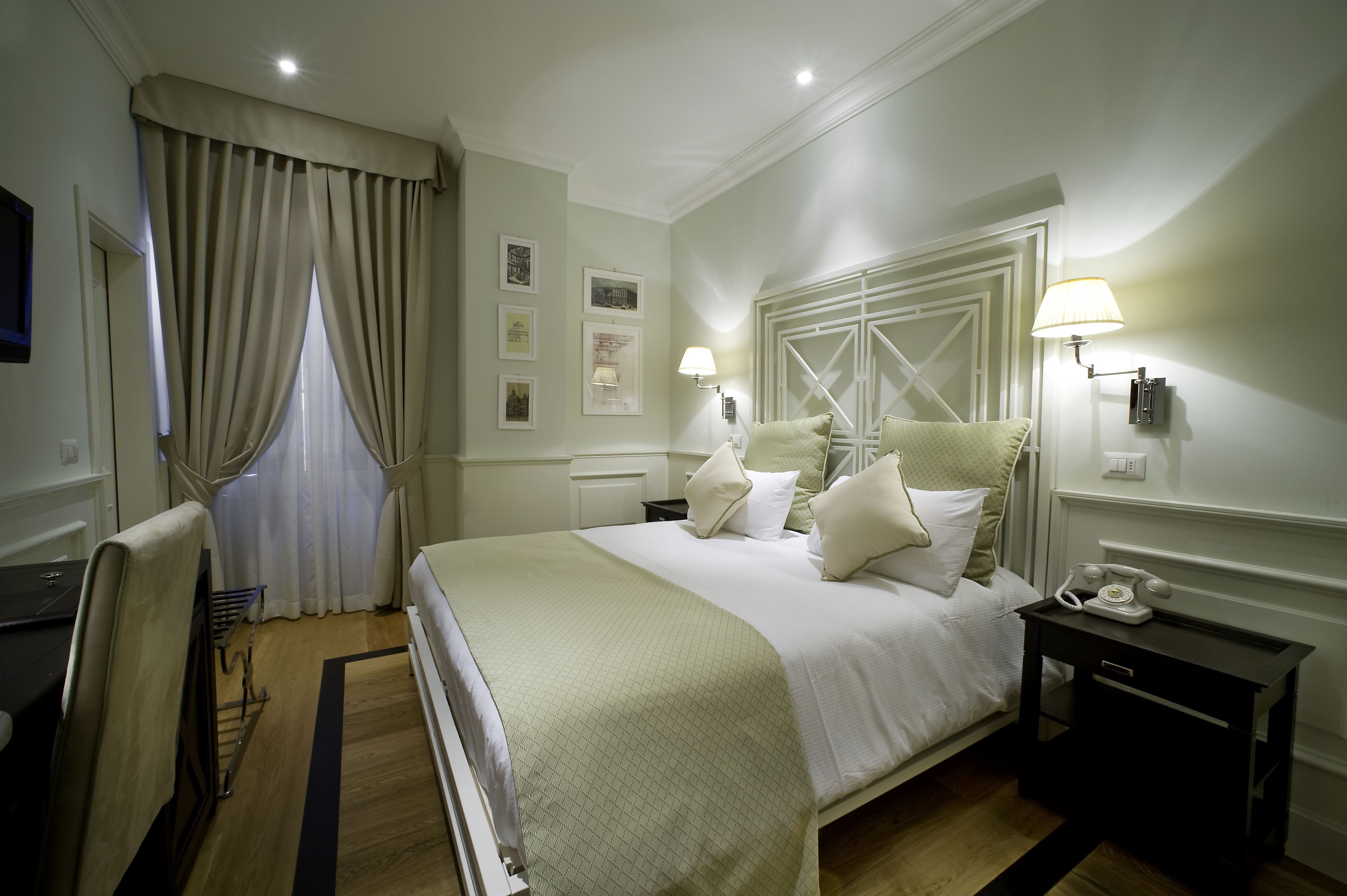 A hotel room with a large bed, two bedside lamps, a nightstand with a telephone, and a window with curtains.