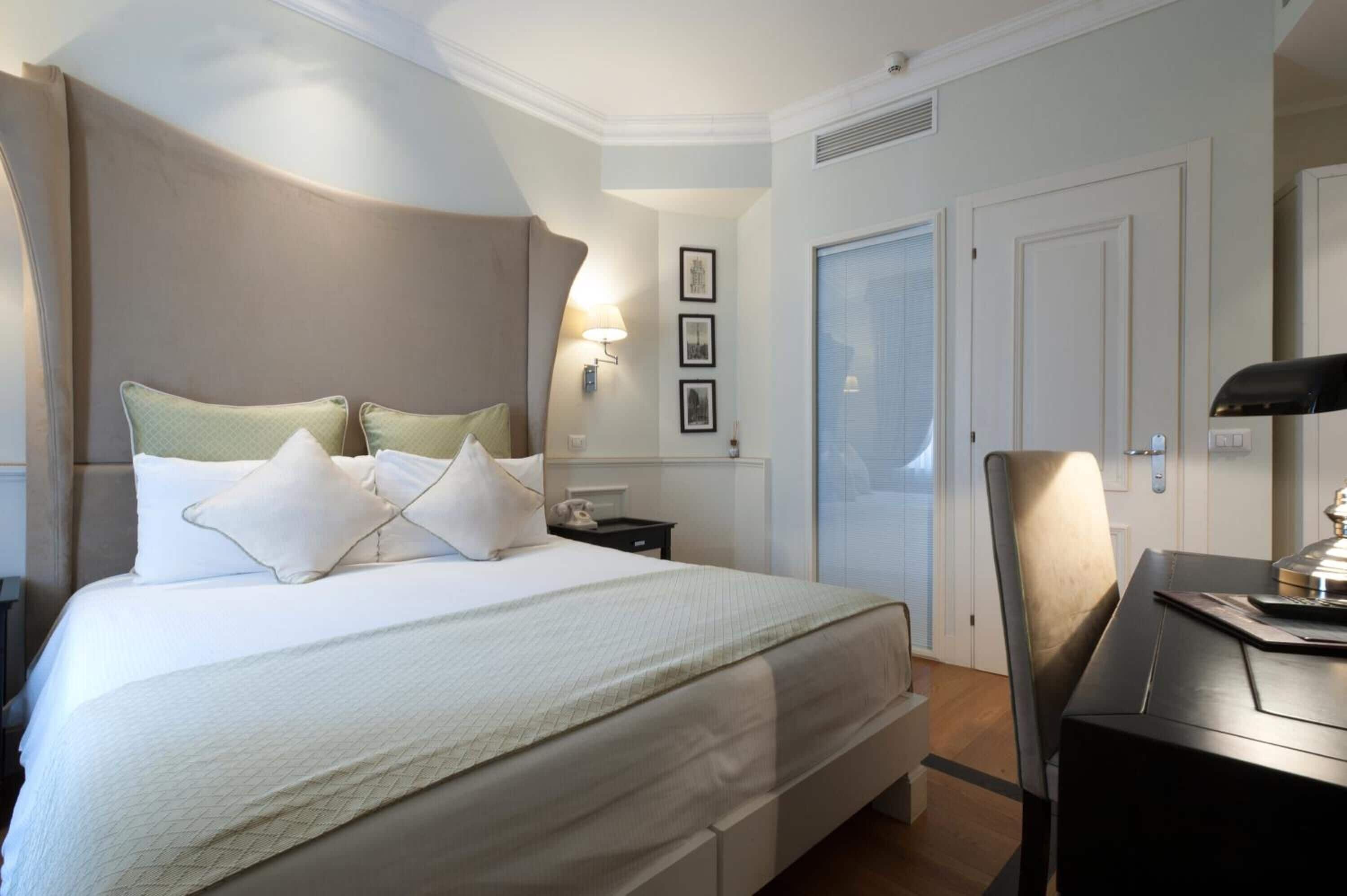 A hotel room with a large bed, bedside tables, a desk, and a chair. There are framed pictures on the wall and a door with a window.