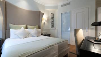 A hotel room with a large bed, bedside tables, a desk, and a chair. There are framed pictures on the wall and a door with a window.