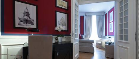 A modern living room with a red accent wall, a dining area with a glass table, and a view into a bedroom with a bed and curtains.