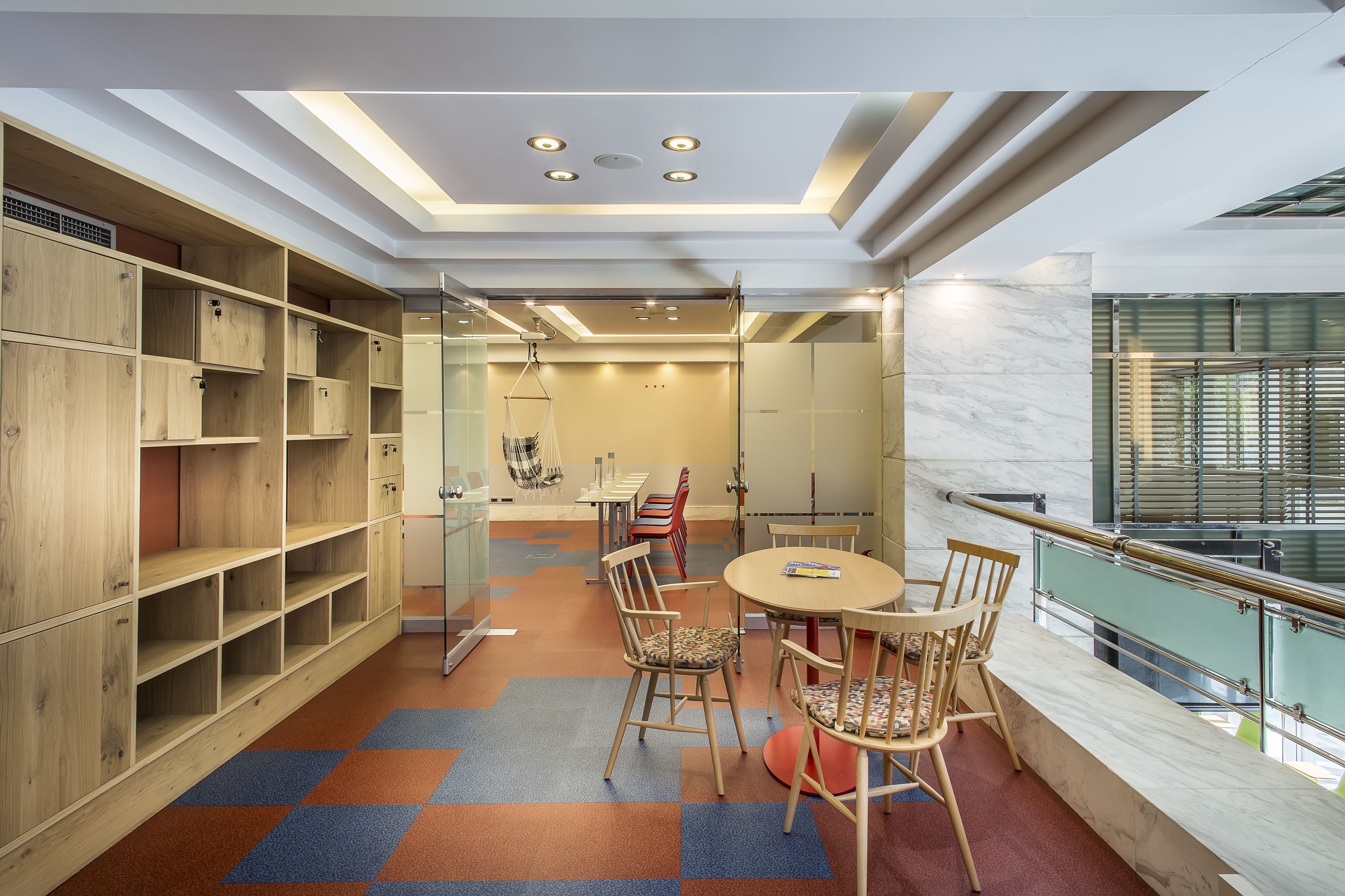 A modern library with wooden bookshelves, a round table with chairs, and a glass partition.