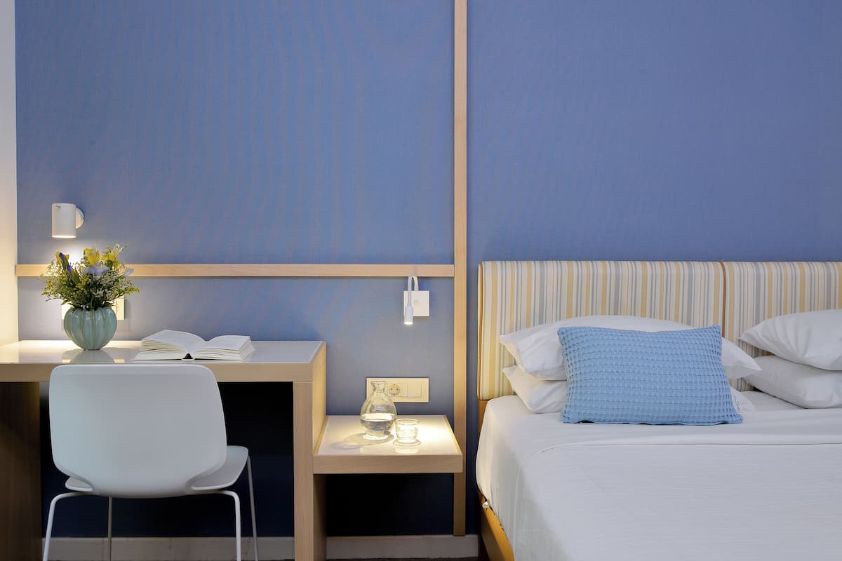 A hotel room with a bed, a bedside table with a lamp, a chair, and a desk with a vase of flowers.