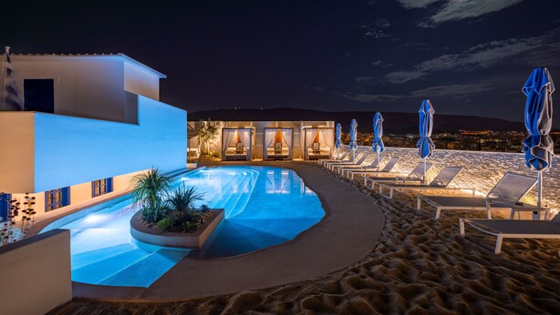 A pool area with lounge chairs and umbrellas, a building with illuminated windows, and a view of the beach at night. - Airotel Alexandros Hotel (Athens)