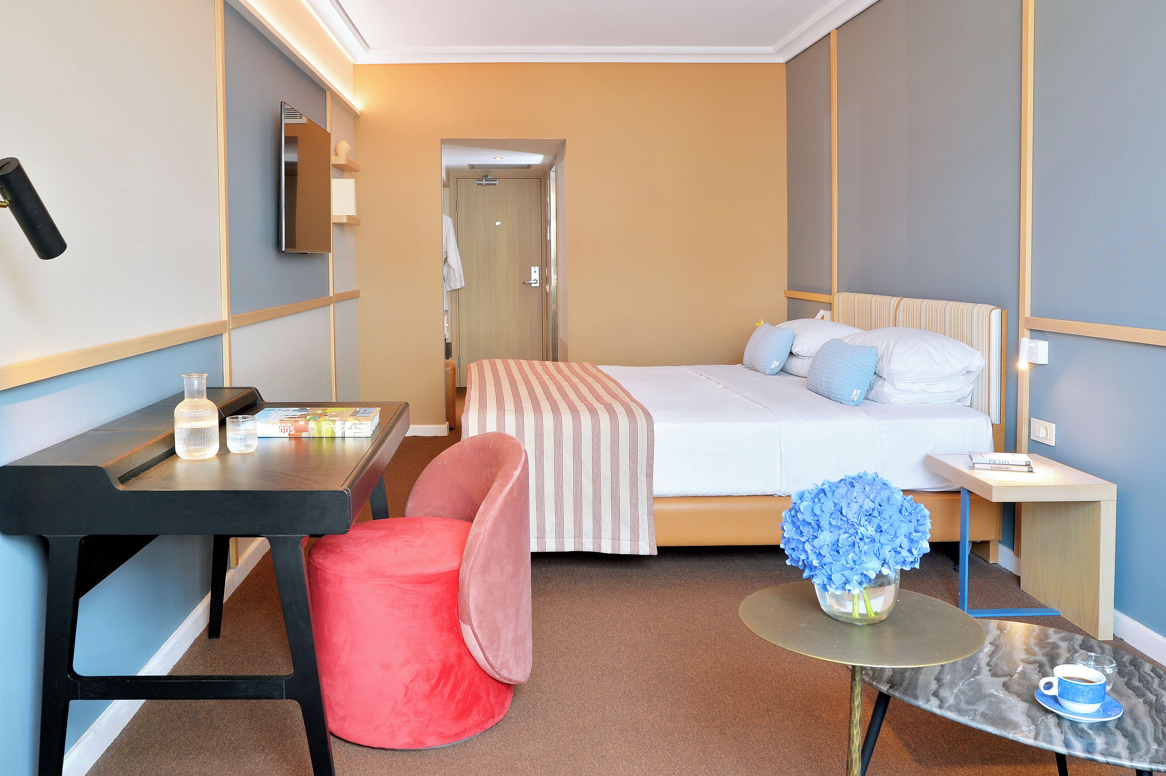 Habitación de hotel con cama, escritorio, silla, una mesita con un florero y una mesita de noche con una taza y platillo.