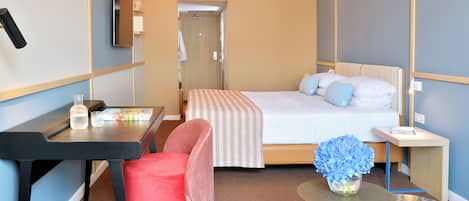 Habitación de hotel con cama, escritorio, silla, una mesita con un florero y una mesita de noche con una taza y platillo.