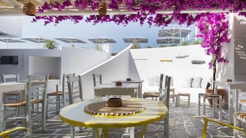 A sunlit outdoor seating area with white furniture and purple flowers, featuring a patterned floor and umbrellas in the background.