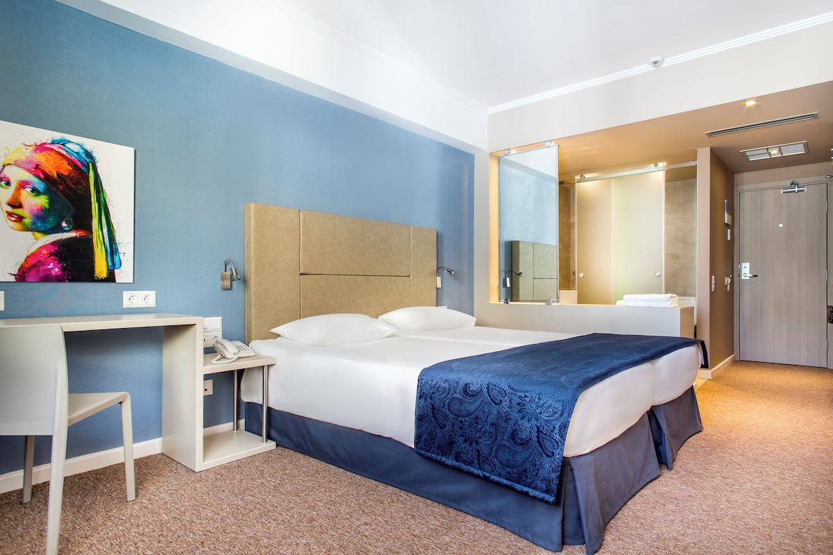 A hotel room with a bed, a desk, a chair, a bathroom with a mirror, and a painting on the wall.