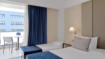 A hotel room with two beds, a small table with a chair, a lamp, and a view of a building outside.