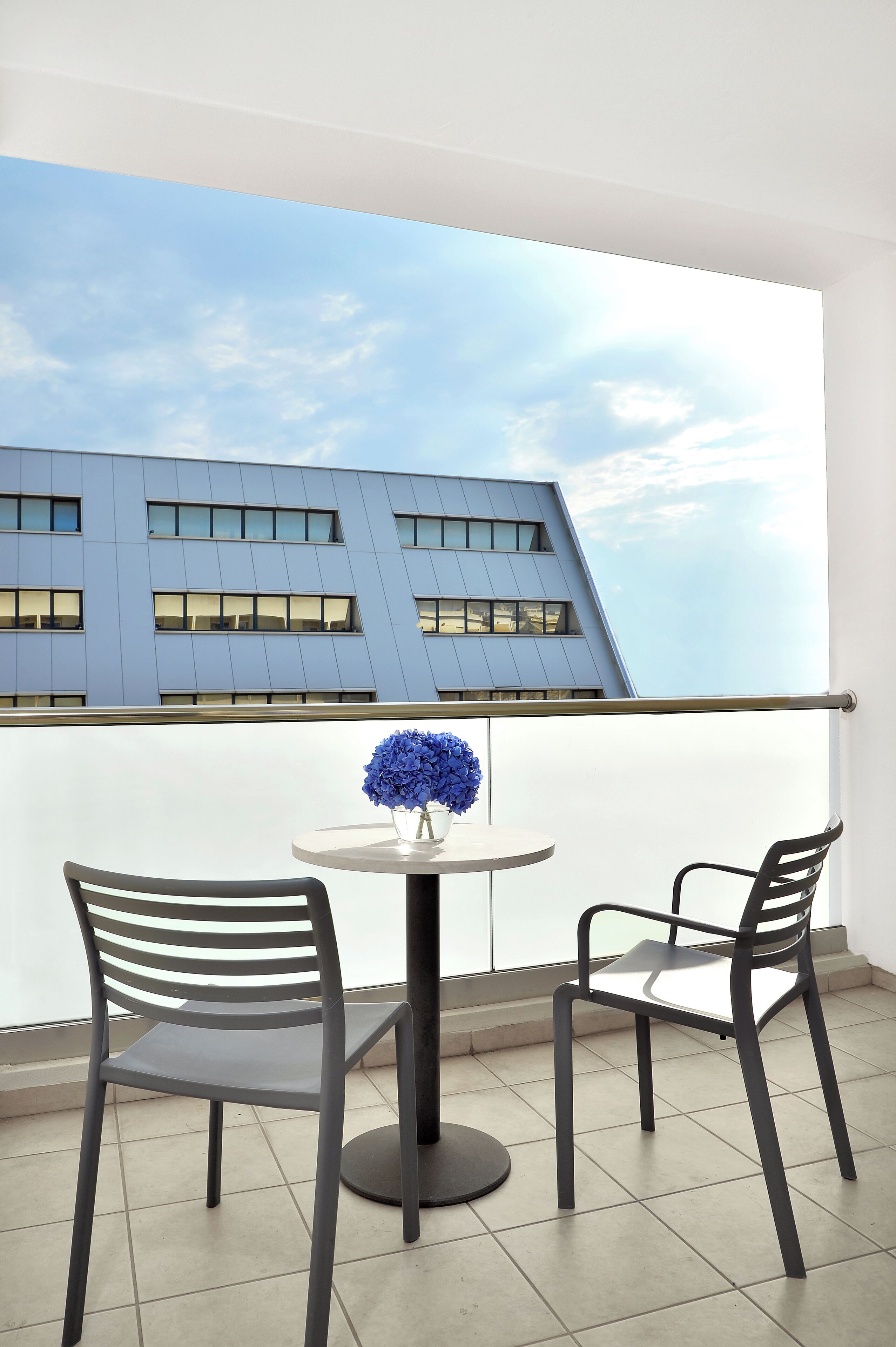 A balcony with two chairs and a table, a vase of flowers, and a view of a building with a modern design.