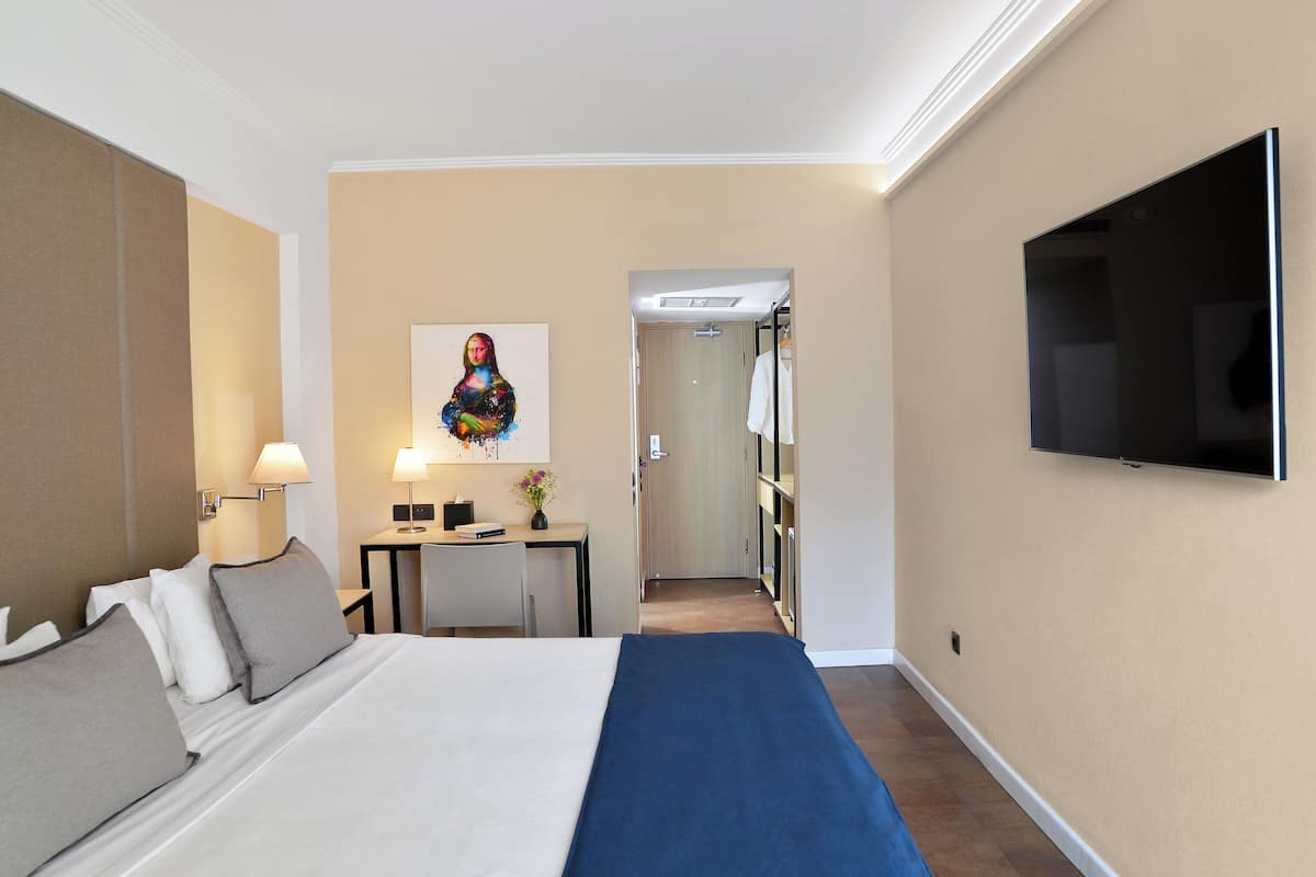 A hotel room with a bed, bedside table, lamp, TV, and a painting on the wall.