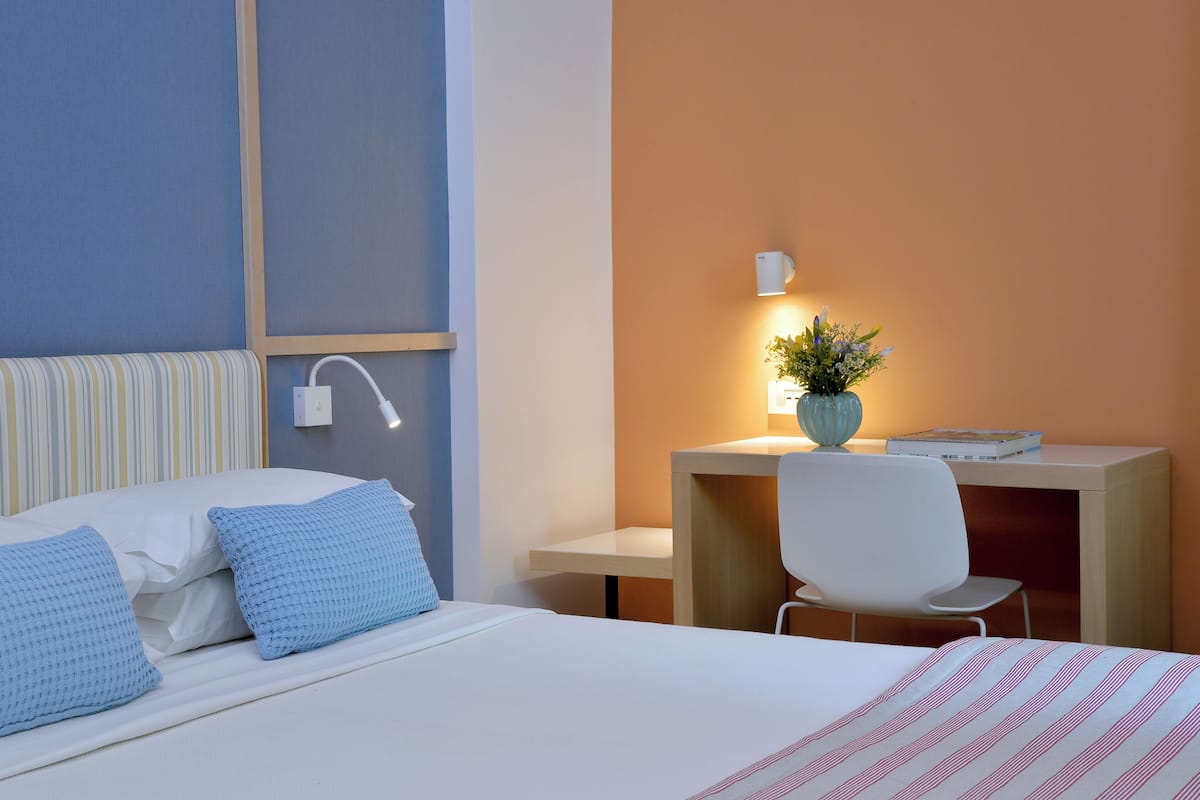 A hotel room with a bed, a desk, a chair, a bedside table, and a wall-mounted lamp.