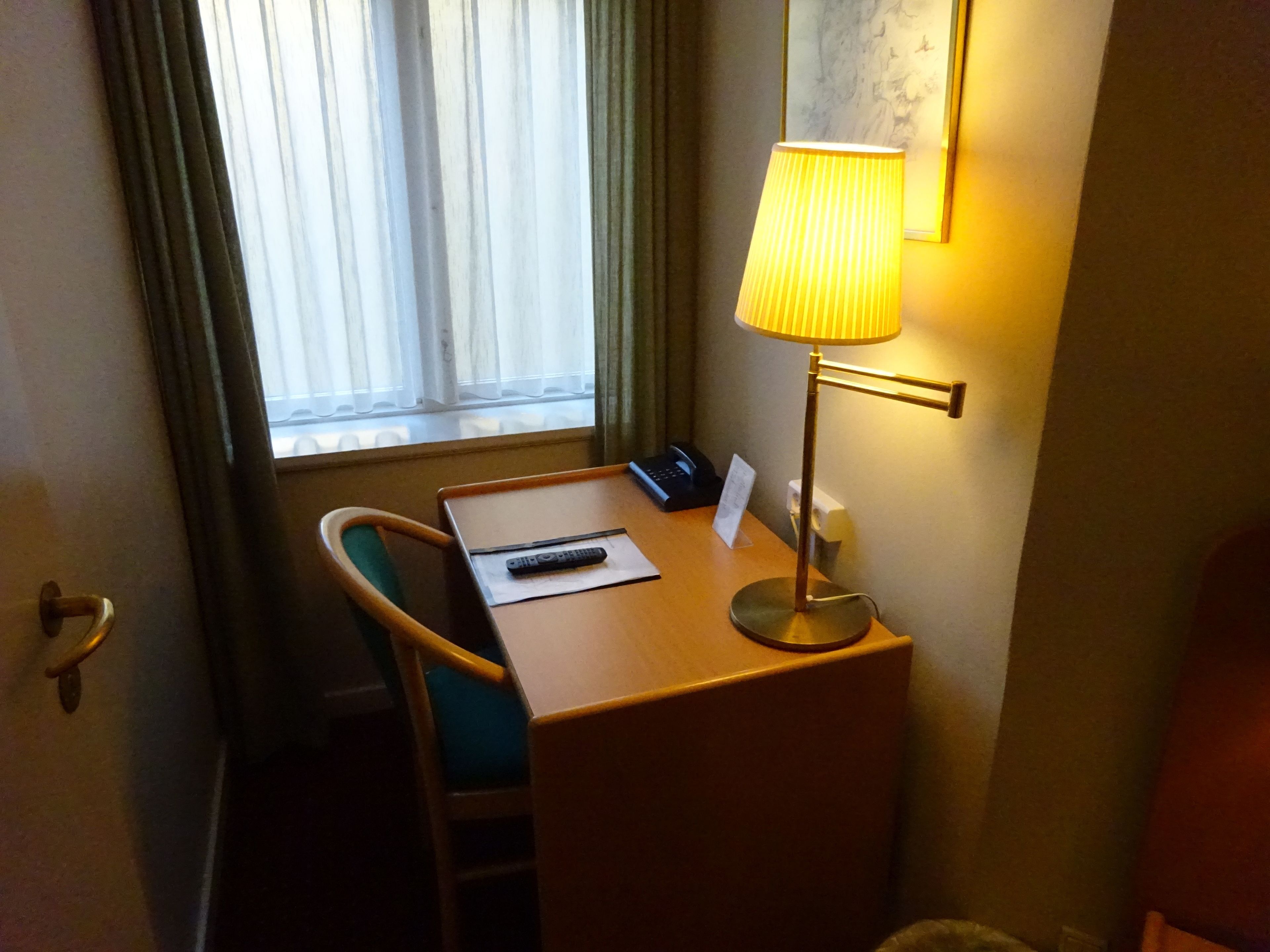 Habitación de hotel con un escritorio de madera, una silla, una lámpara, un control remoto y una ventana con cortinas.