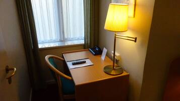 Habitación de hotel con un escritorio de madera, una silla, una lámpara, un control remoto y una ventana con cortinas.