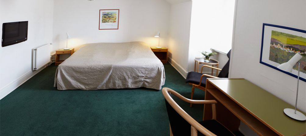 Et hotelværelse med seng, skrivebord, stol, lampe og billeder på væggene.