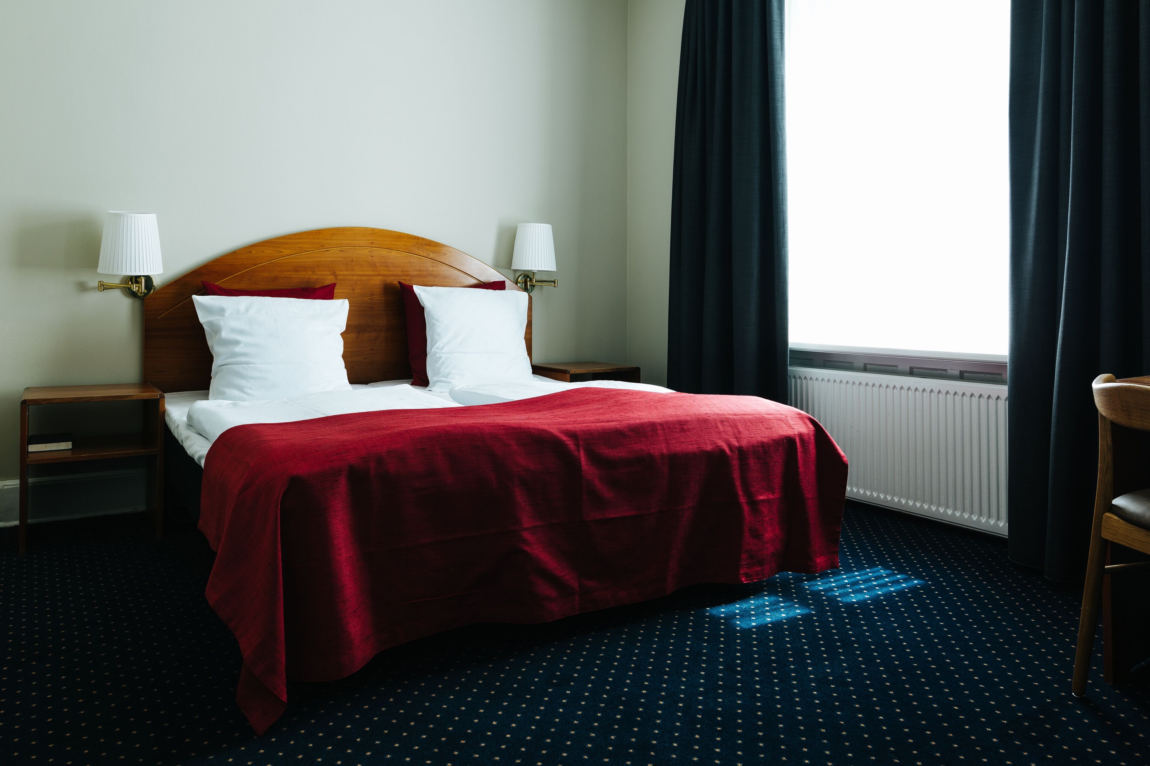 Habitación de hotel con una cama, dos lámparas de noche, una ventana con cortinas y un radiador.