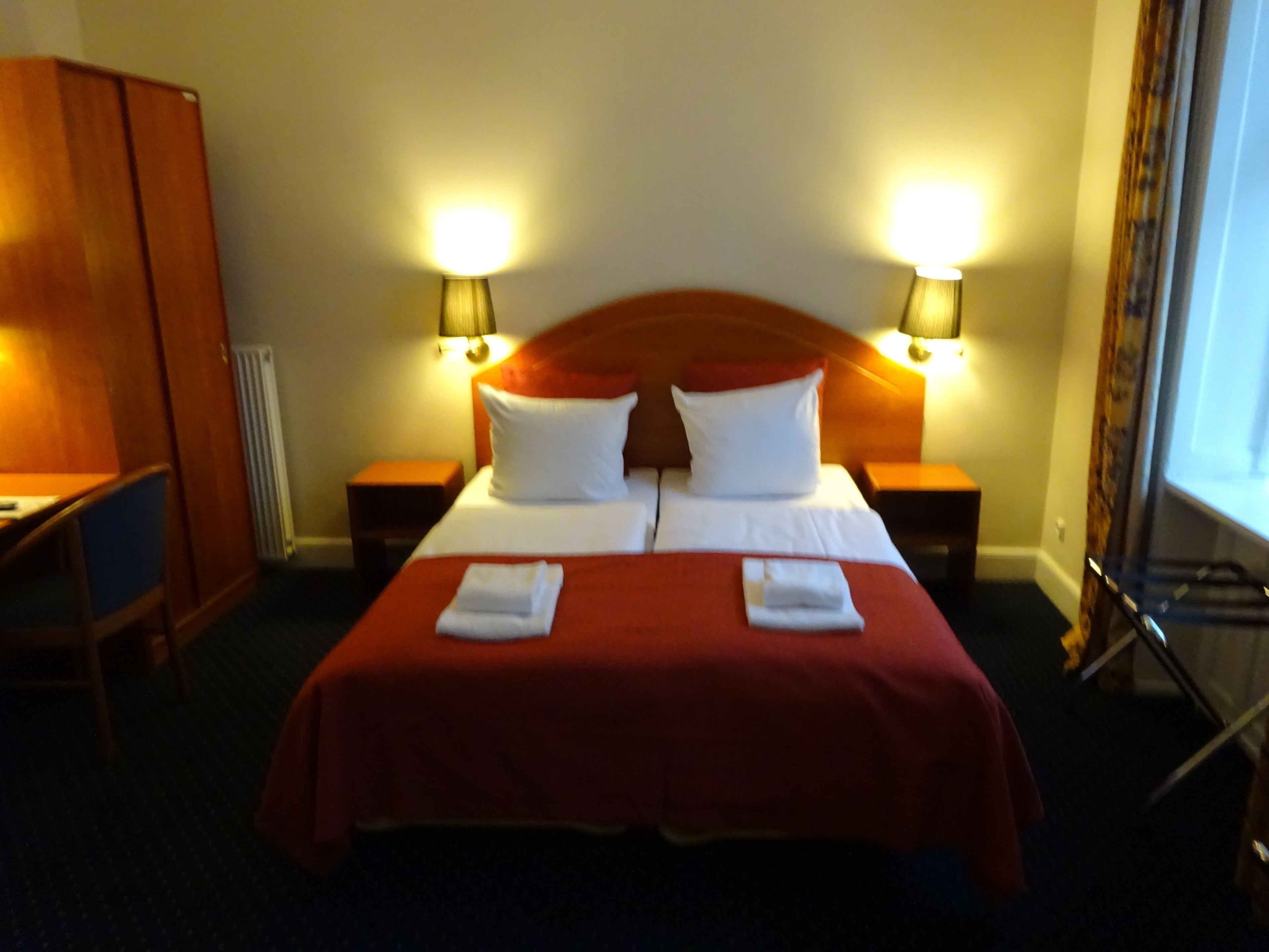 Quarto de hotel com uma cama grande, duas mesas de cabeceira, um guarda-roupa de madeira e duas luminárias fixadas na parede.