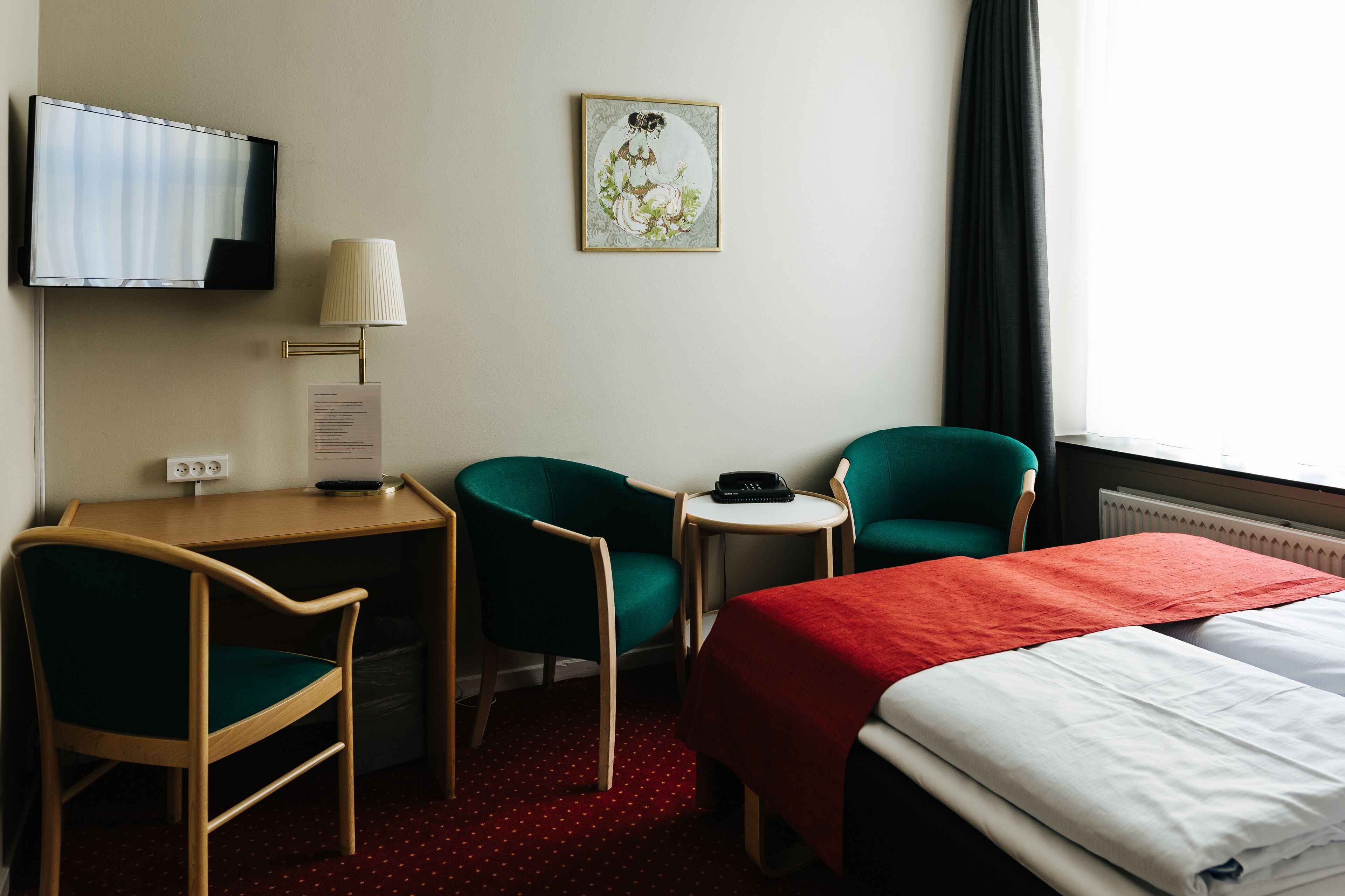 Ein Hotelzimmer mit Schreibtisch, zwei Stühlen, einem Bett, einem Fernseher, einer Lampe und einem gerahmten Bild an der Wand.