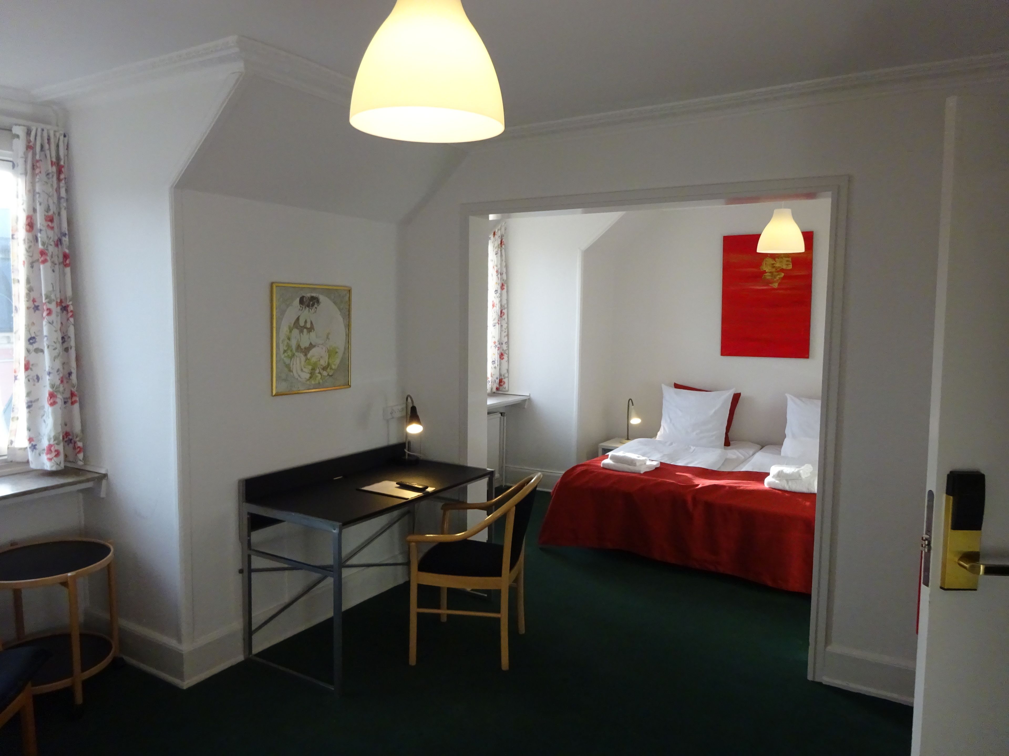 A hotel room with a desk, chair, bed, and a red bedspread.