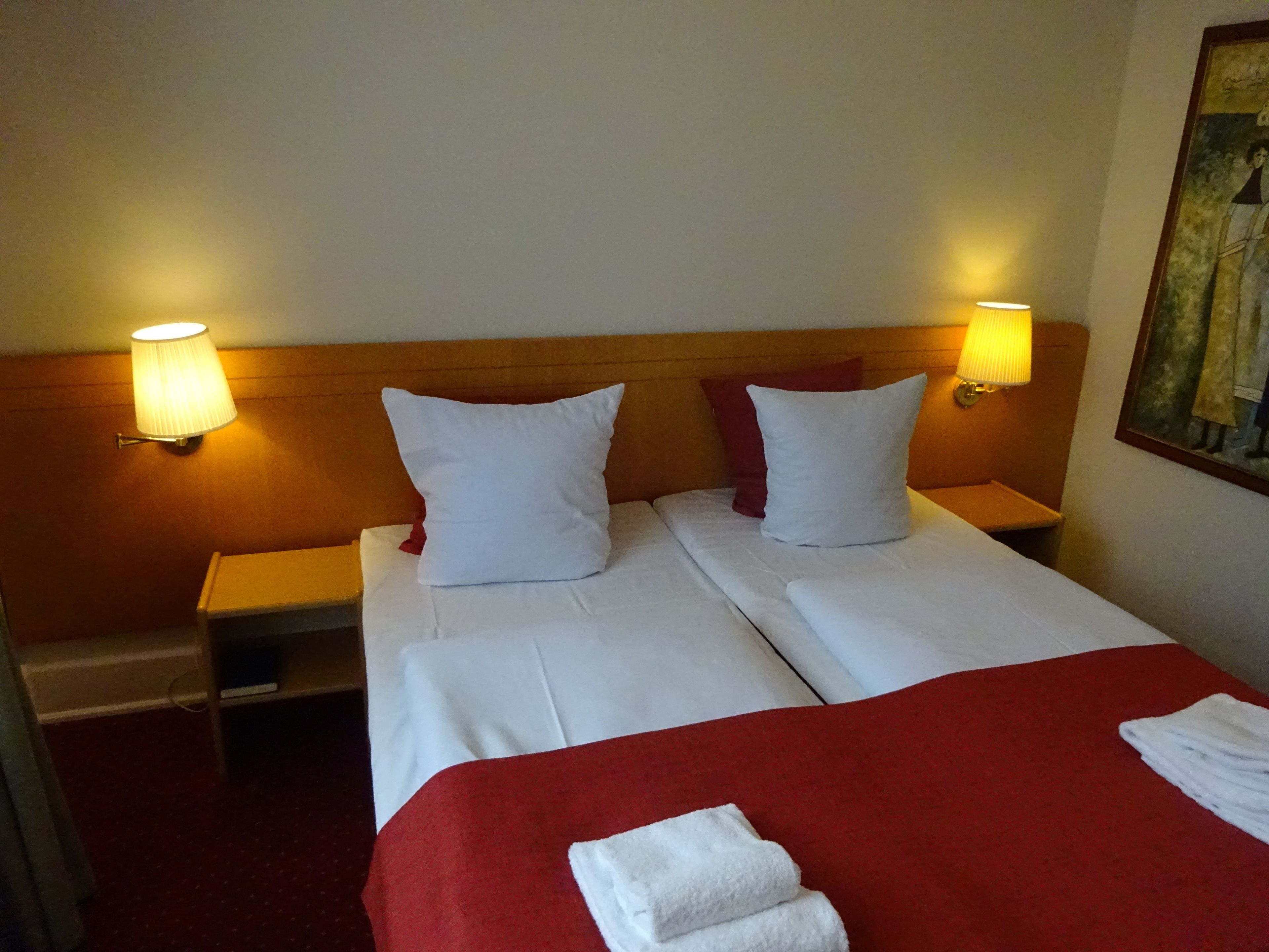 Une chambre d’hôtel avec deux lits, chacun étant équipé de draps et d’oreillers blancs, d’une couverture rouge et d’une serviette pliée posée sur le lit. Deux lampes murales sont fixées de chaque côté des lits.