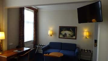 Habitación de hotel con un sofá azul, un escritorio con lámpara, un cuadro en la pared y un televisor de pantalla plana montado en la pared.
