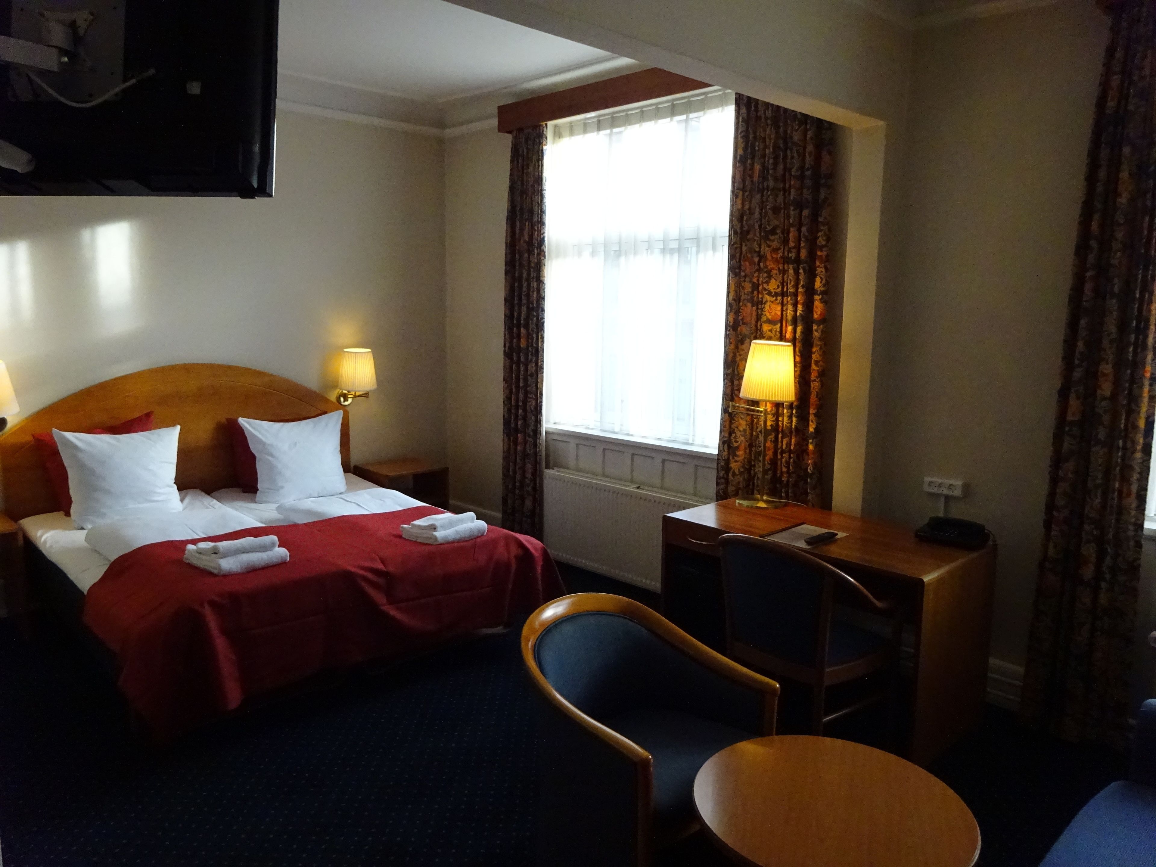 A hotel room with a bed, two bedside lamps, a desk with a chair, and a view of the outdoors through a window.