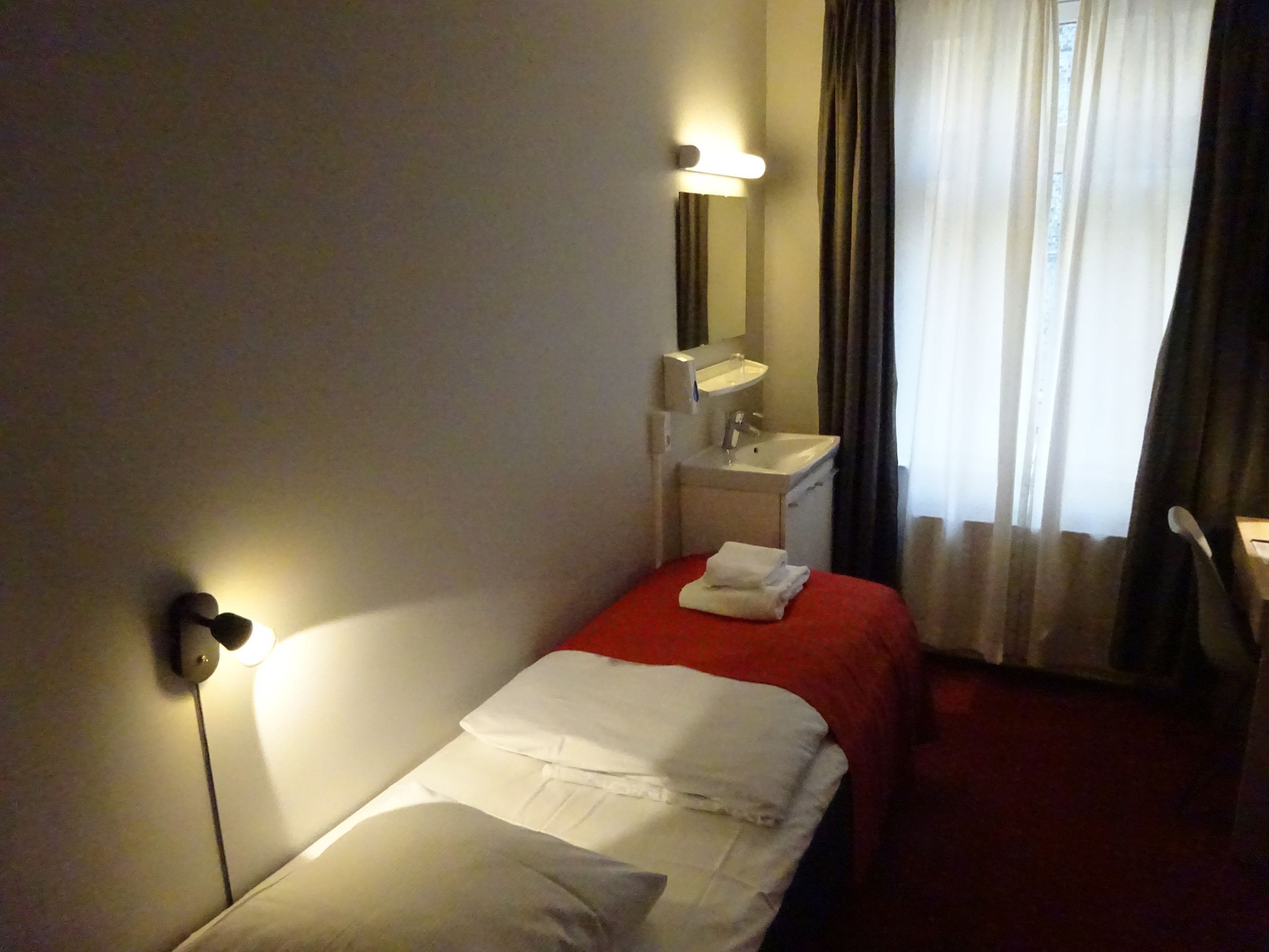 A hotel room with a single bed, a bedside table with a lamp, a mirror, a small sink, and a window with curtains.