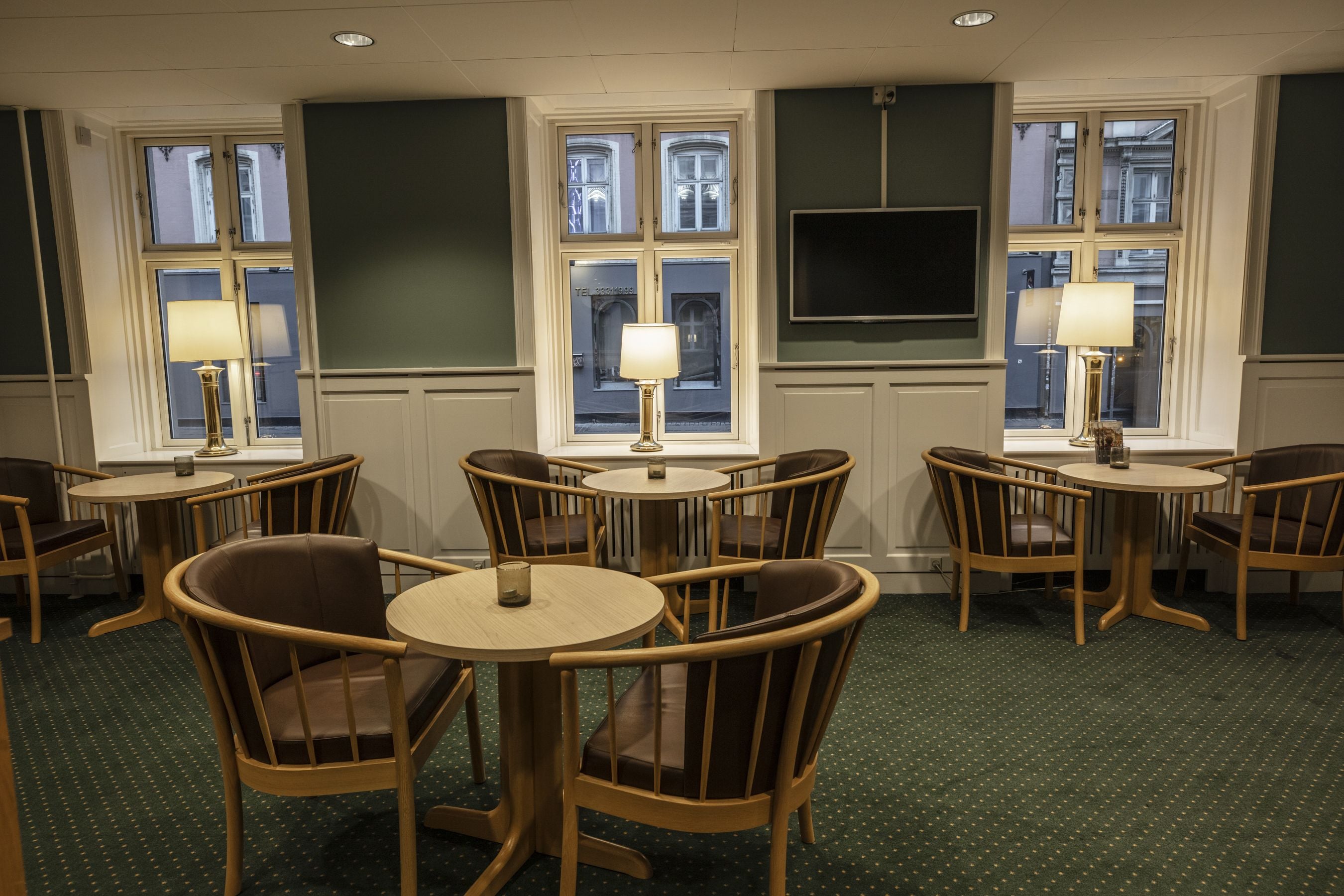 A hotel room with a green carpet, wooden chairs, round tables, and a TV mounted on the wall.