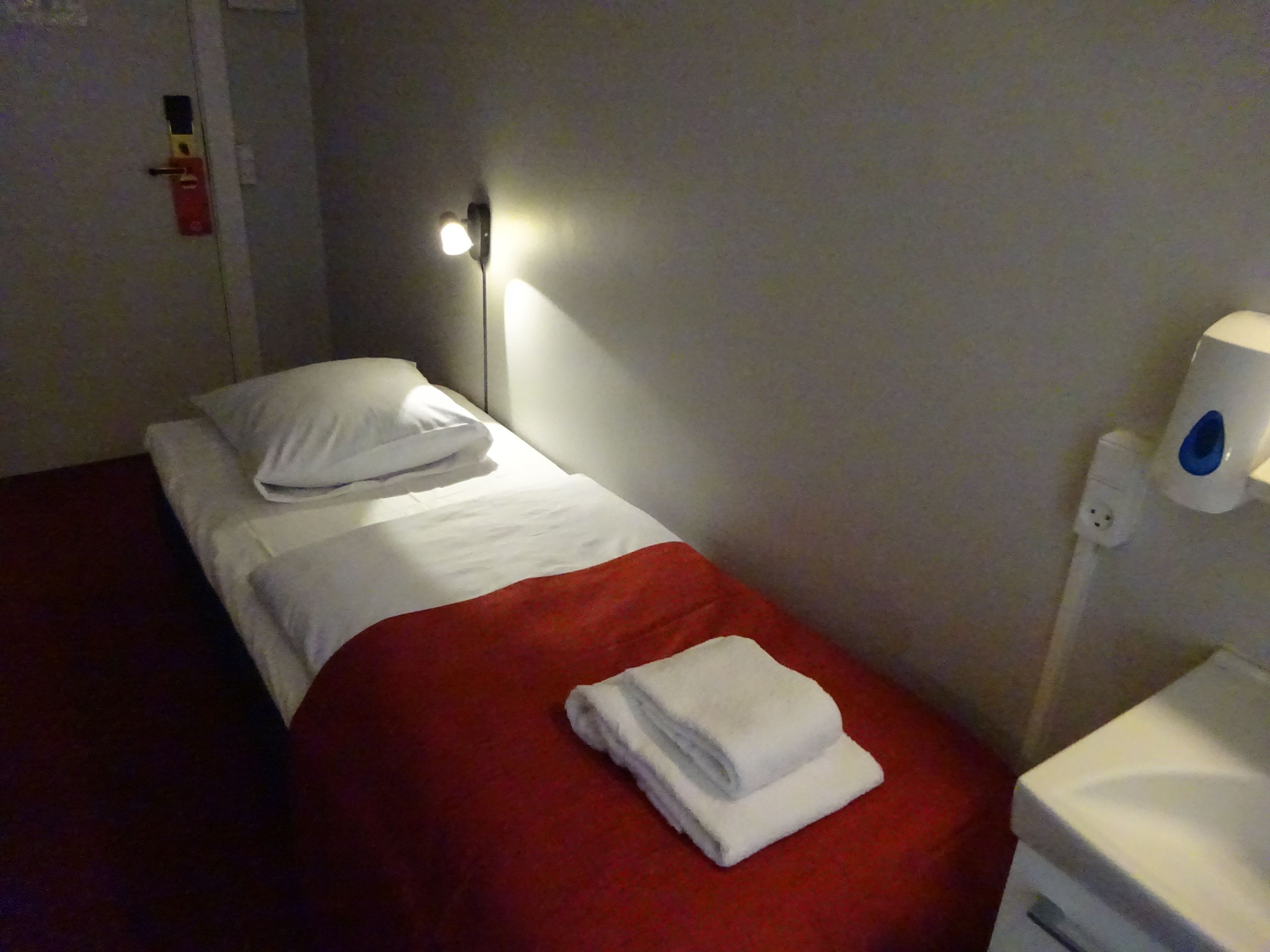 Uma cama de solteiro com roupa de cama branca e manta vermelha, uma toalha branca na cama, uma luz noturna fixada na parede e uma tomada branca com um plugue.