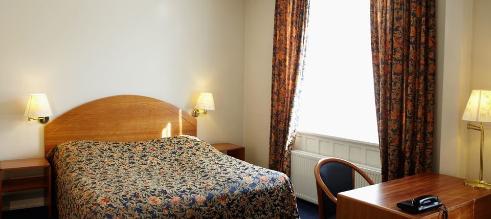 Una camera d'albergo con un letto, due lampade sui comodini, una scrivania con il telefono e delle tende.