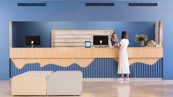 Een receptiebalie met een blauwe wand, twee monitoren, een tablet en een plant. Voor de balie staan twee poefs.