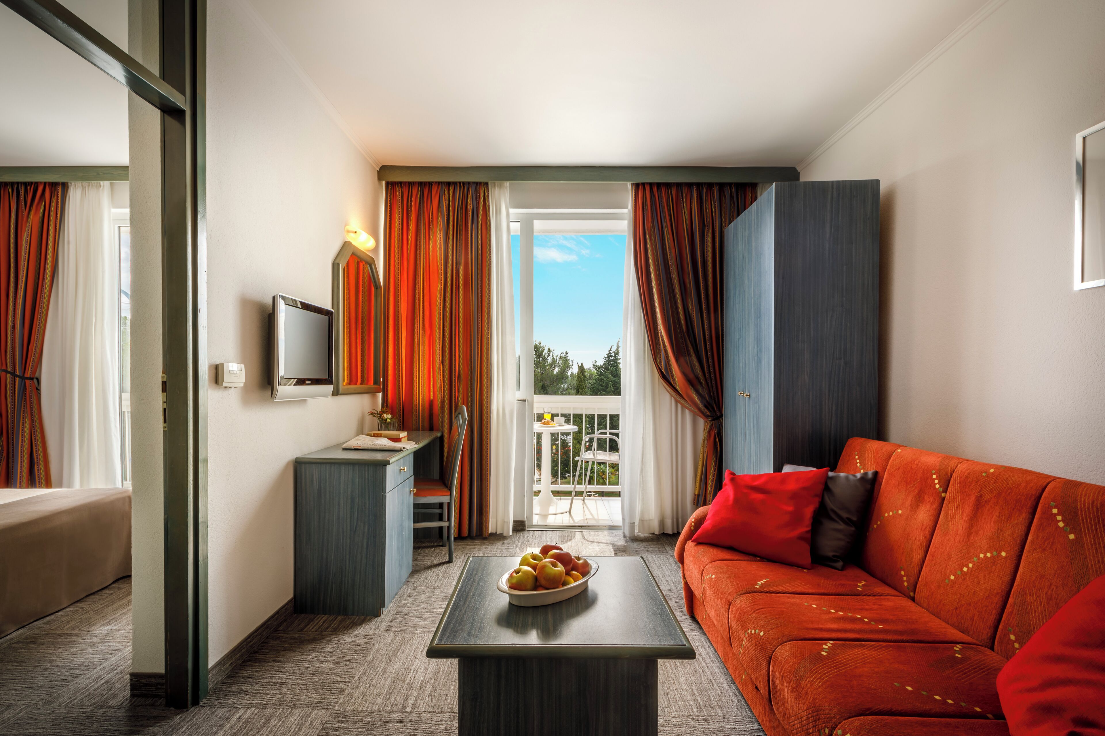 A hotel room with a sofa, a small table with fruit, a television, and a balcony with curtains.
