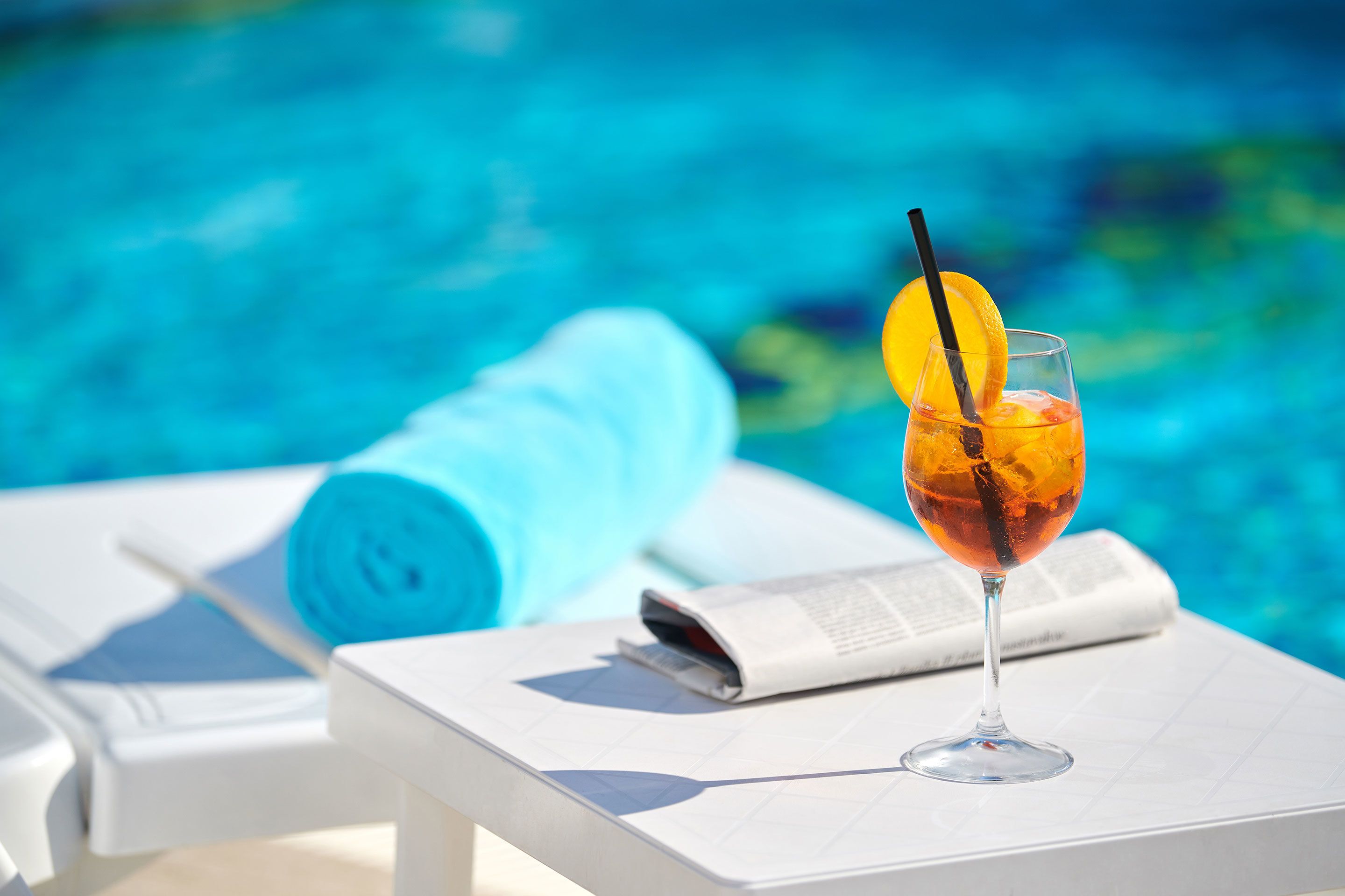 A glass of iced tea with a lemon slice and straw on a white table by a pool.