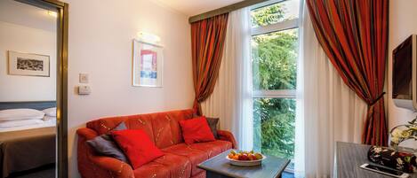 A hotel room with a red sofa, a small table with fruit, a bed, and a window with curtains.