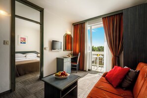 A hotel room with a bed, a desk with a TV, a dining table with fruit, a sofa with red cushions, and a balcony with curtains. - Laguna Maradiso Hotel by Aminess  (Novigrad)