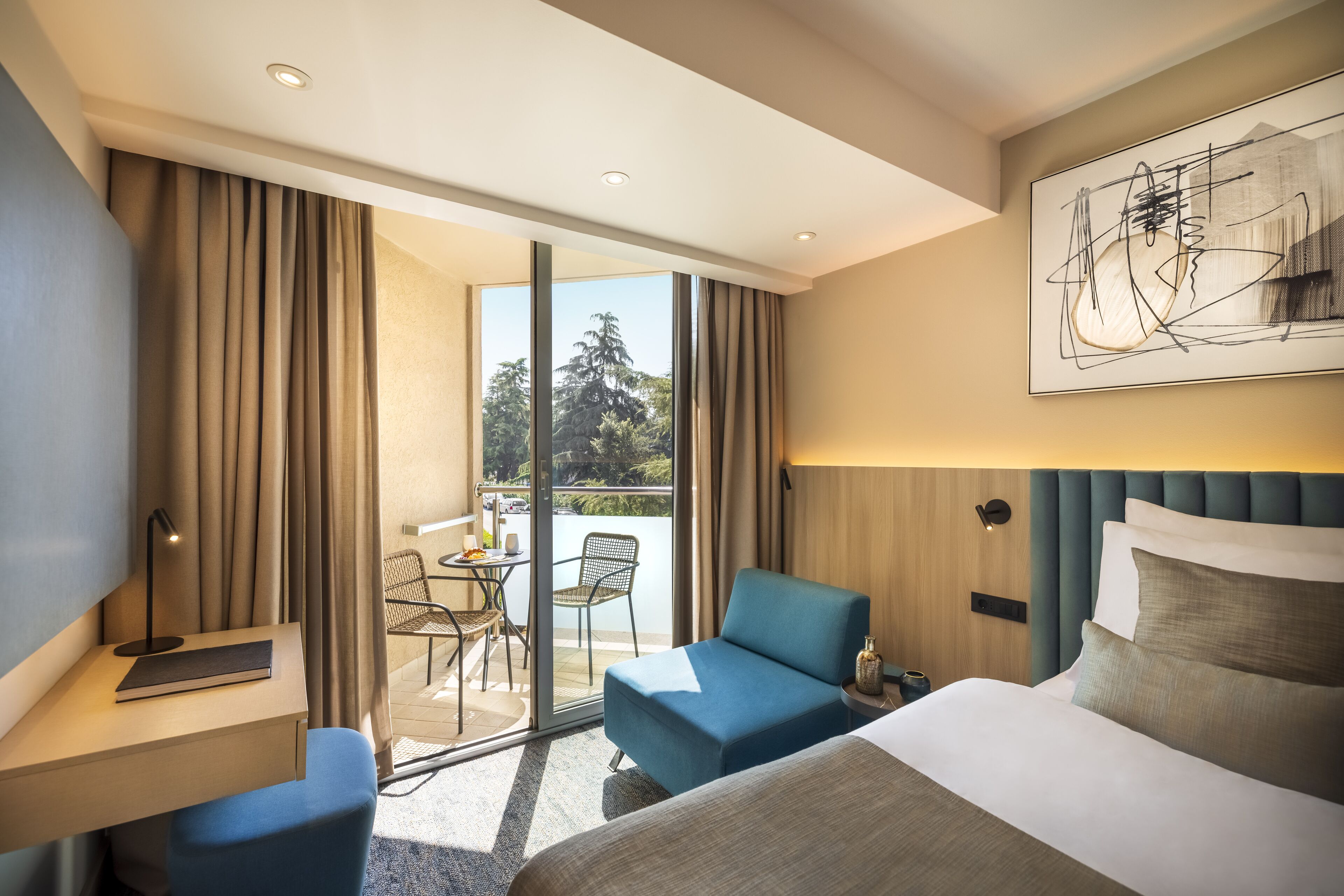 Ein Hotelzimmer mit Bett, Schreibtisch, Stuhl, einem Balkon mit Tisch und Stühlen sowie einem großen Fenster.