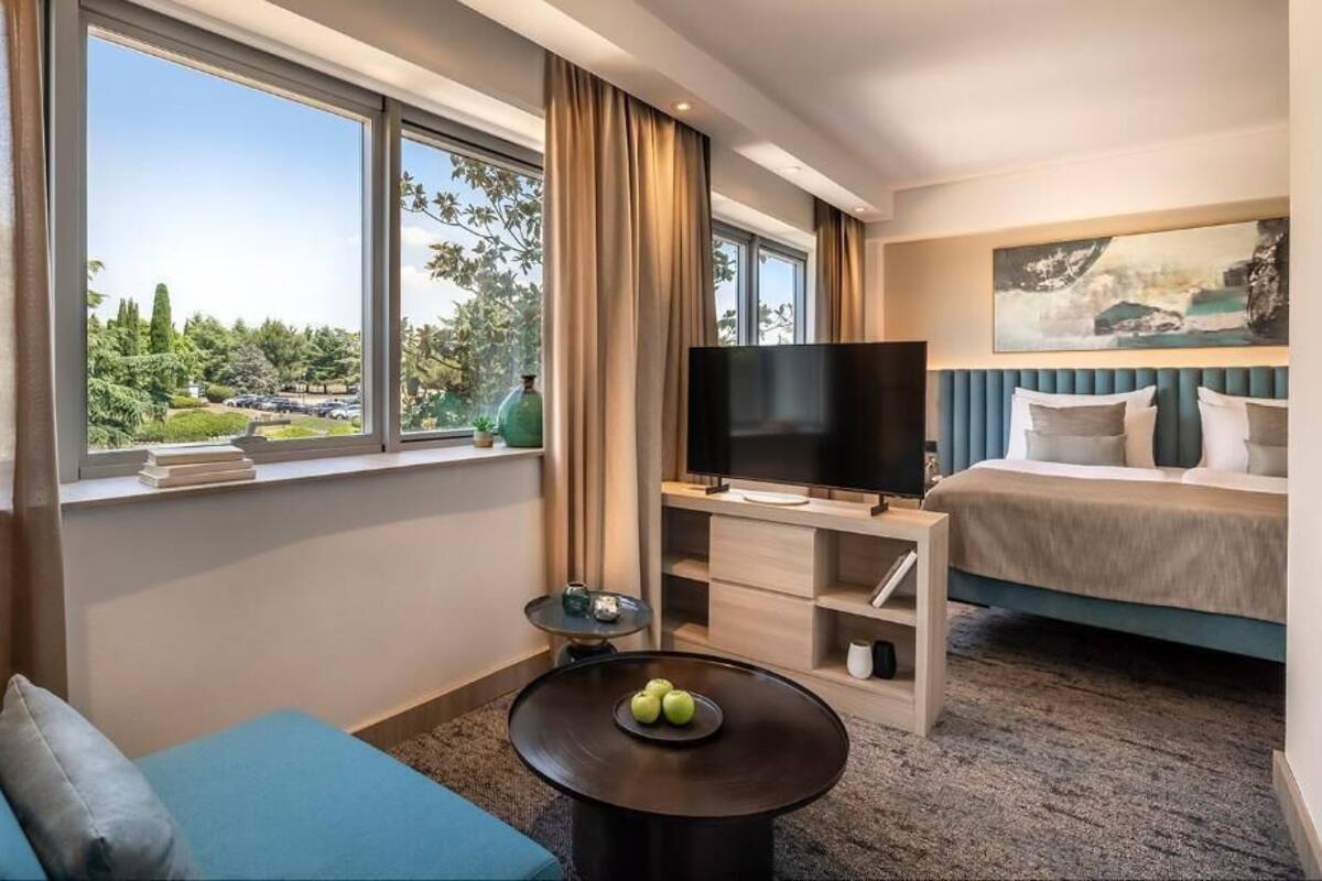 A hotel room with a bed, a TV, a sofa, a coffee table with apples, and a view of greenery outside.